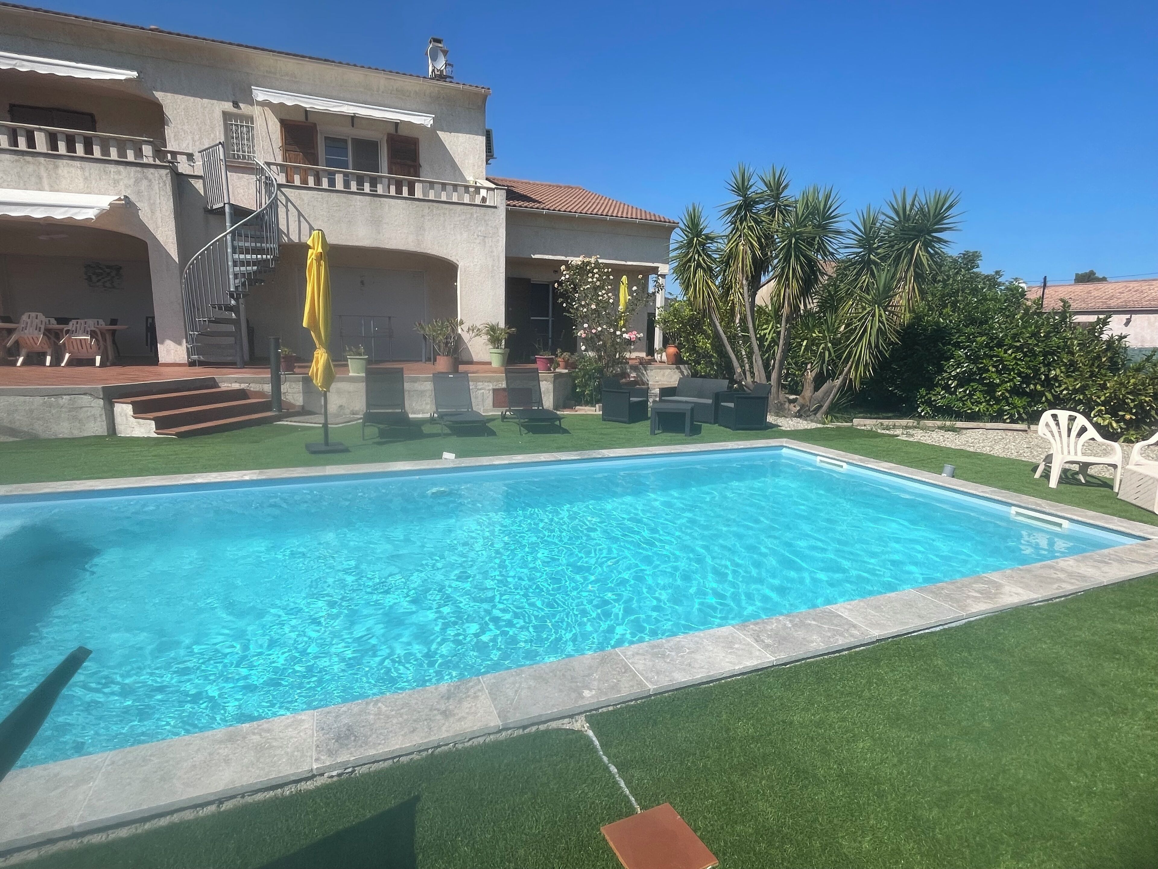 Una piscina con agua cristalina azul, rodeada de césped artificial, y un edificio con balcón y mobiliario de exterior.