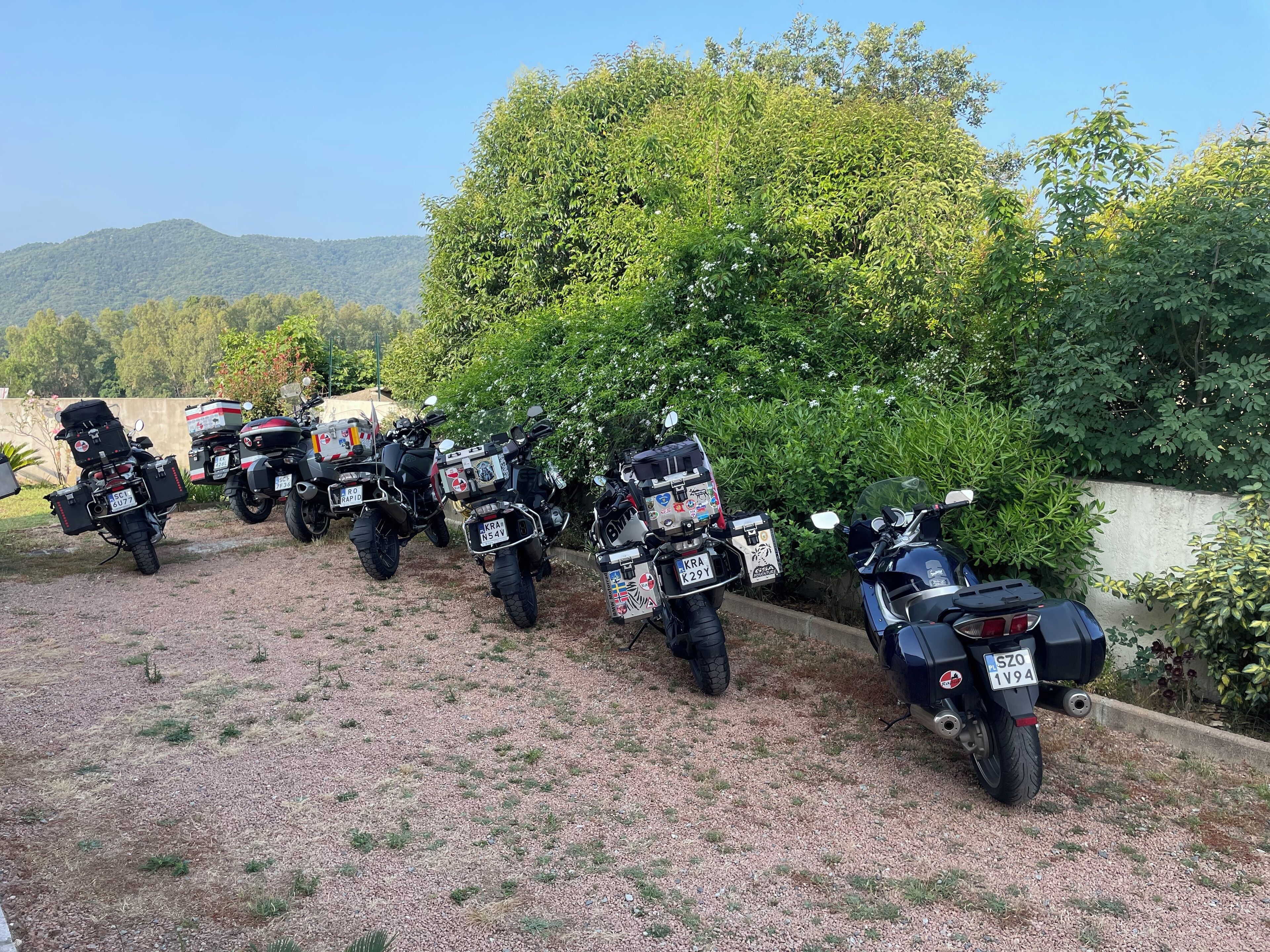 Una hilera de motocicletas estacionadas sobre un sendero de grava, con un fondo de vegetación y colinas.