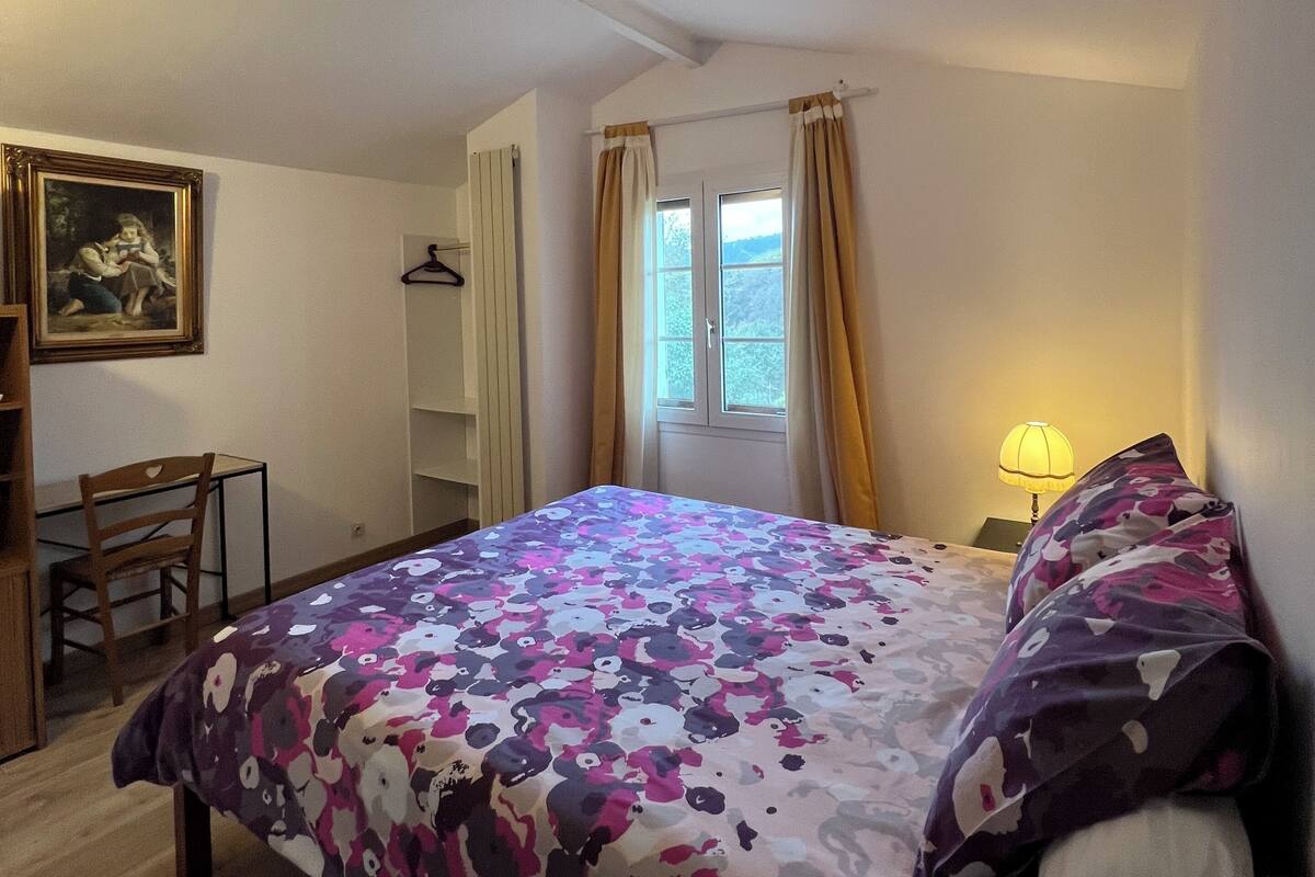 Un dormitorio con cama, escritorio, silla, ventana con cortinas y lámpara.