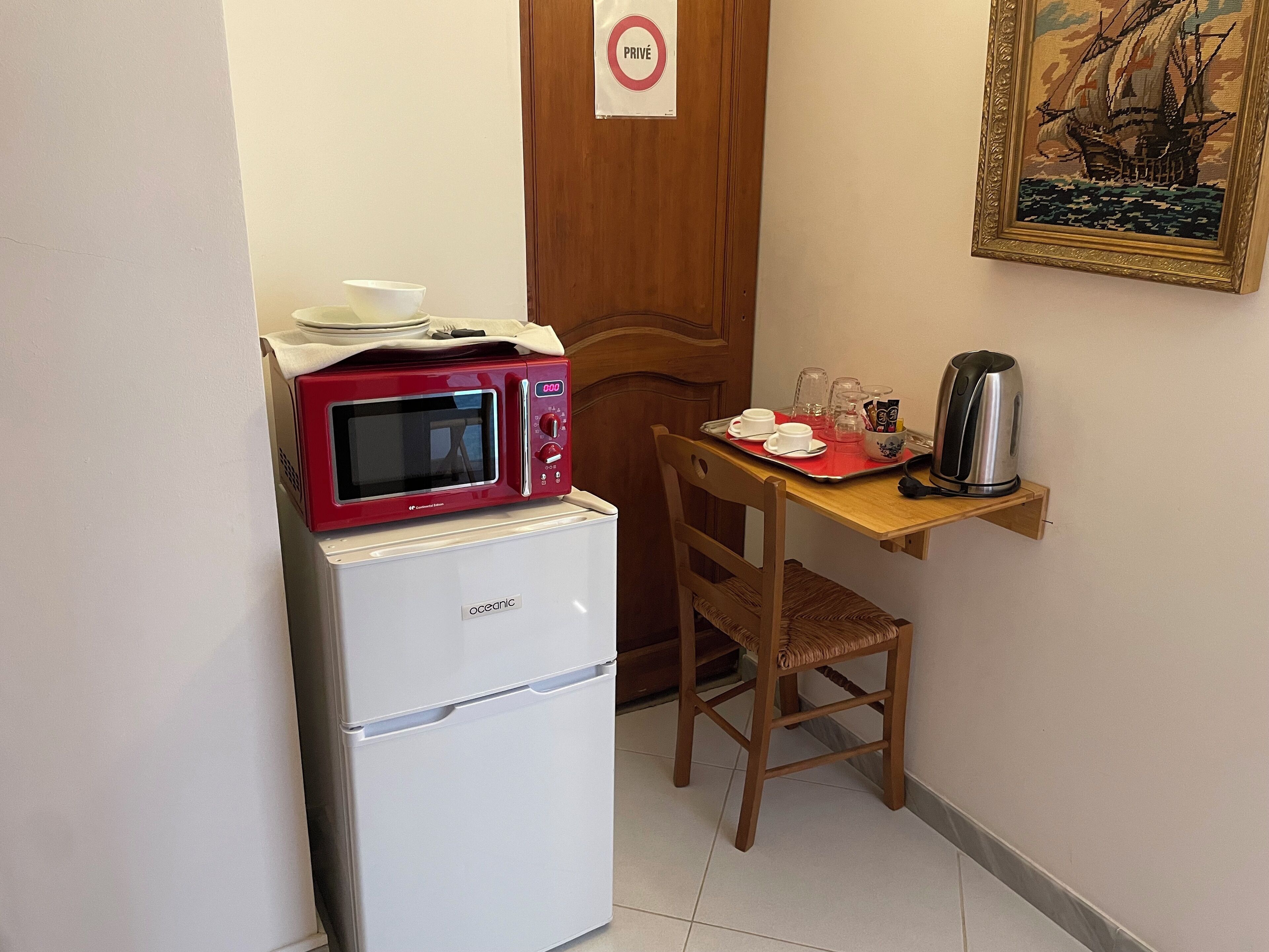 Een kleine keuken met een rode magnetron bovenop een witte koelkast, een houten stoel, een klein tafeltje met een dienblad met bekers en een waterkoker, en een lijstje aan de muur.