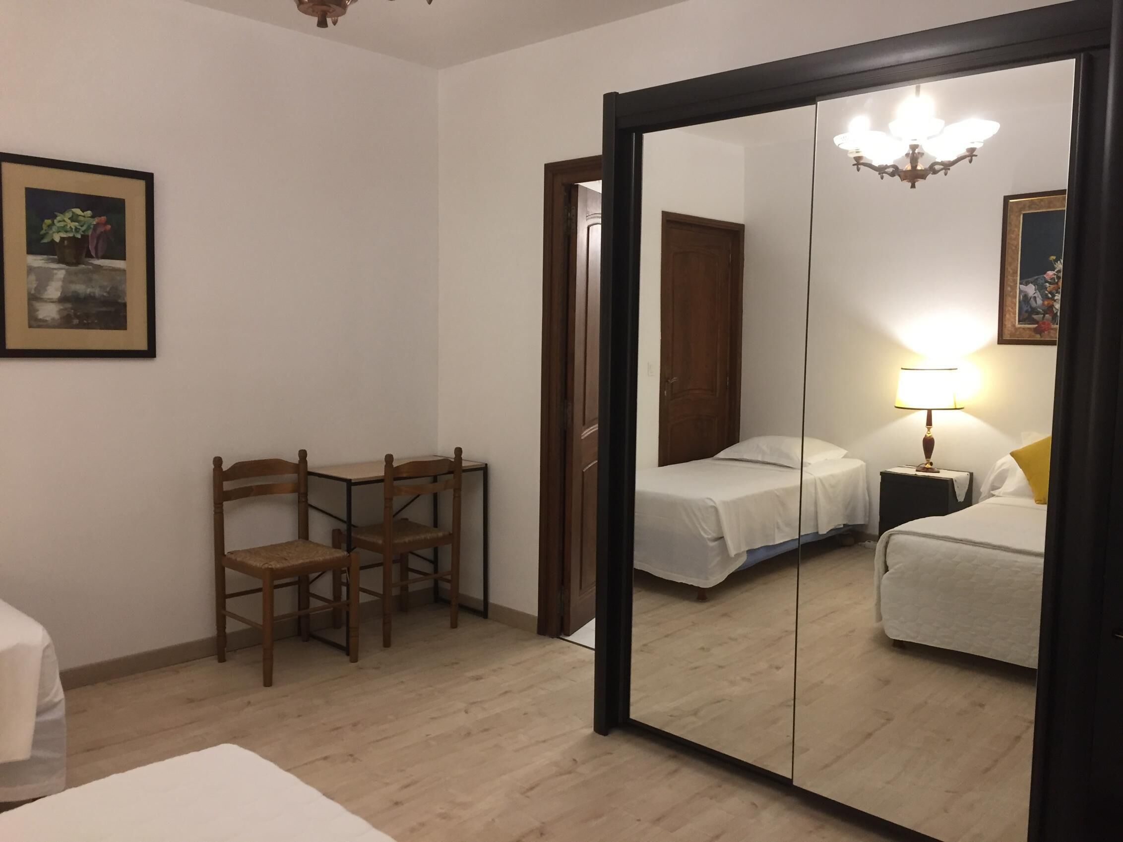 Een slaapkamer met een houten stoel, een bed, een nachtkastje met een lamp en een spiegel die de kamer weerspiegelt.