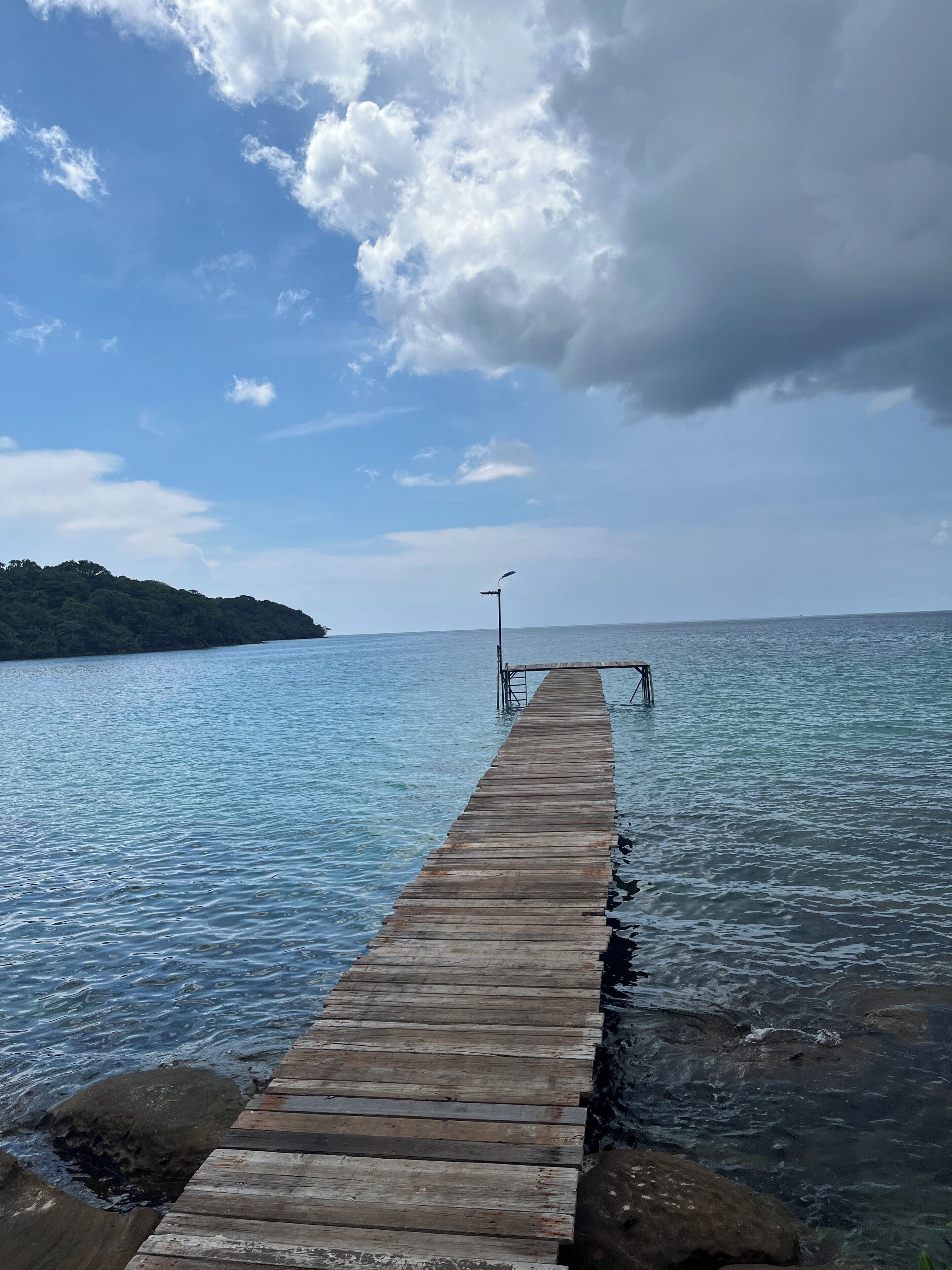 Un muelle de madera se adentra en aguas tranquilas, bajo un cielo despejado y con una isla cubierta de bosque a lo lejos.