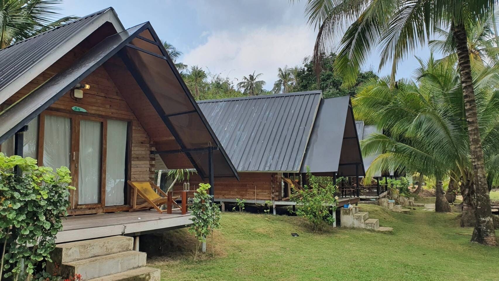 A wooden cabin with a sloped roof, a porch with a chair, and a small garden area with palm trees.