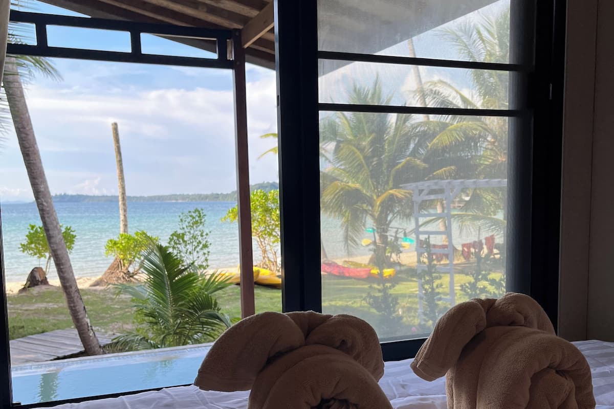 A bed with two towels shaped like animals, a view of the sea and palm trees through large windows, and a wooden ceiling.