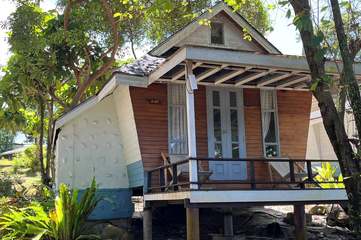 A small, two-story wooden house with a balcony, surrounded by trees and vegetation.