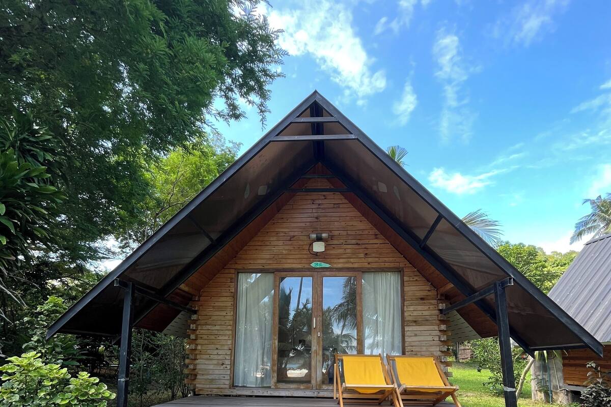 A wooden cabin with a sloped roof, a porch with a yellow chair, and a stone staircase leading to the entrance.