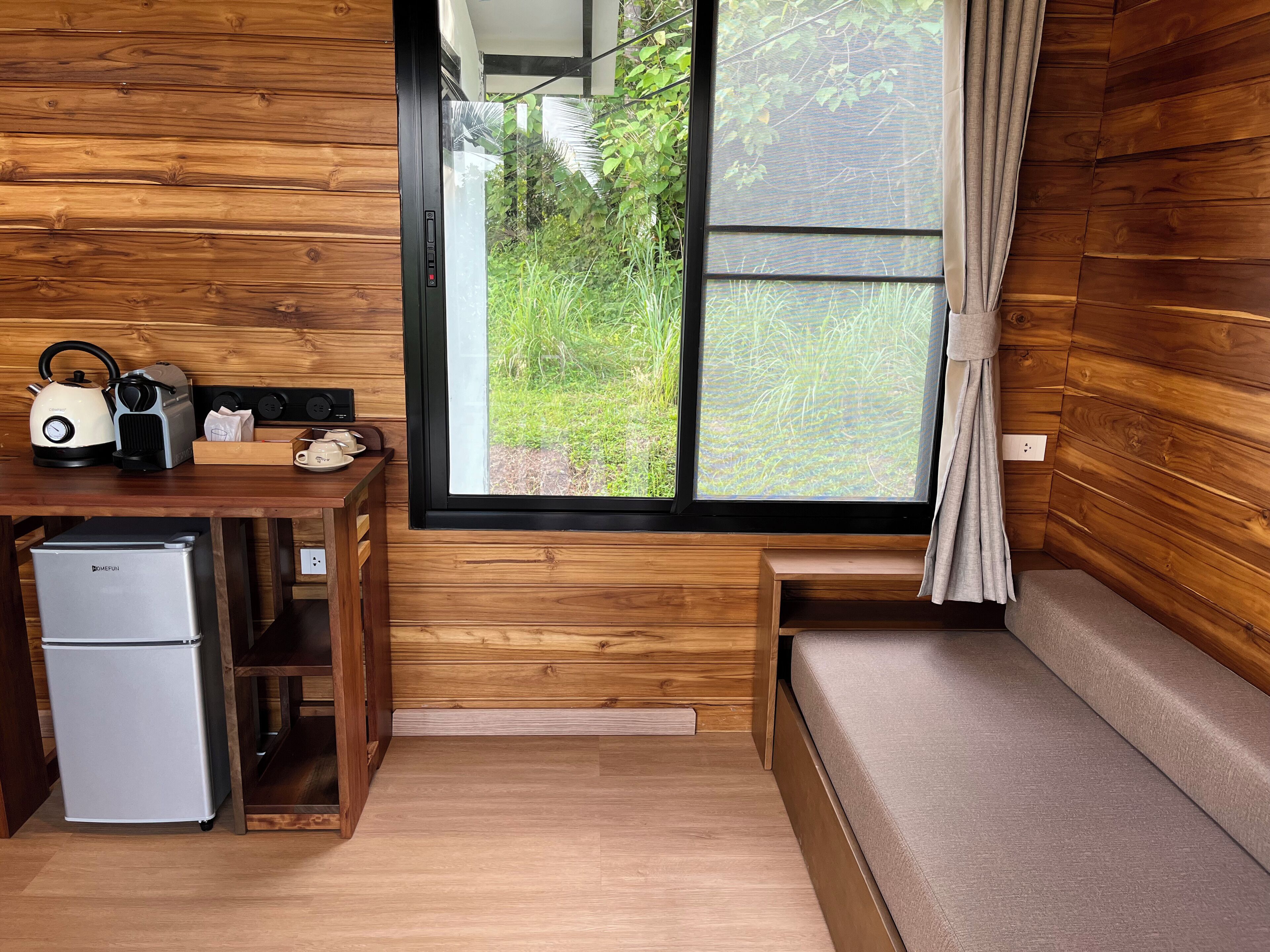 Een knus ingerichte kamer met houten details, een kleine kitchenette, een bank en een raam met uitzicht op groen.
