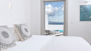 A bedroom with a bed, two patterned pillows, a view of the sea through an open door, and a painting on the wall.