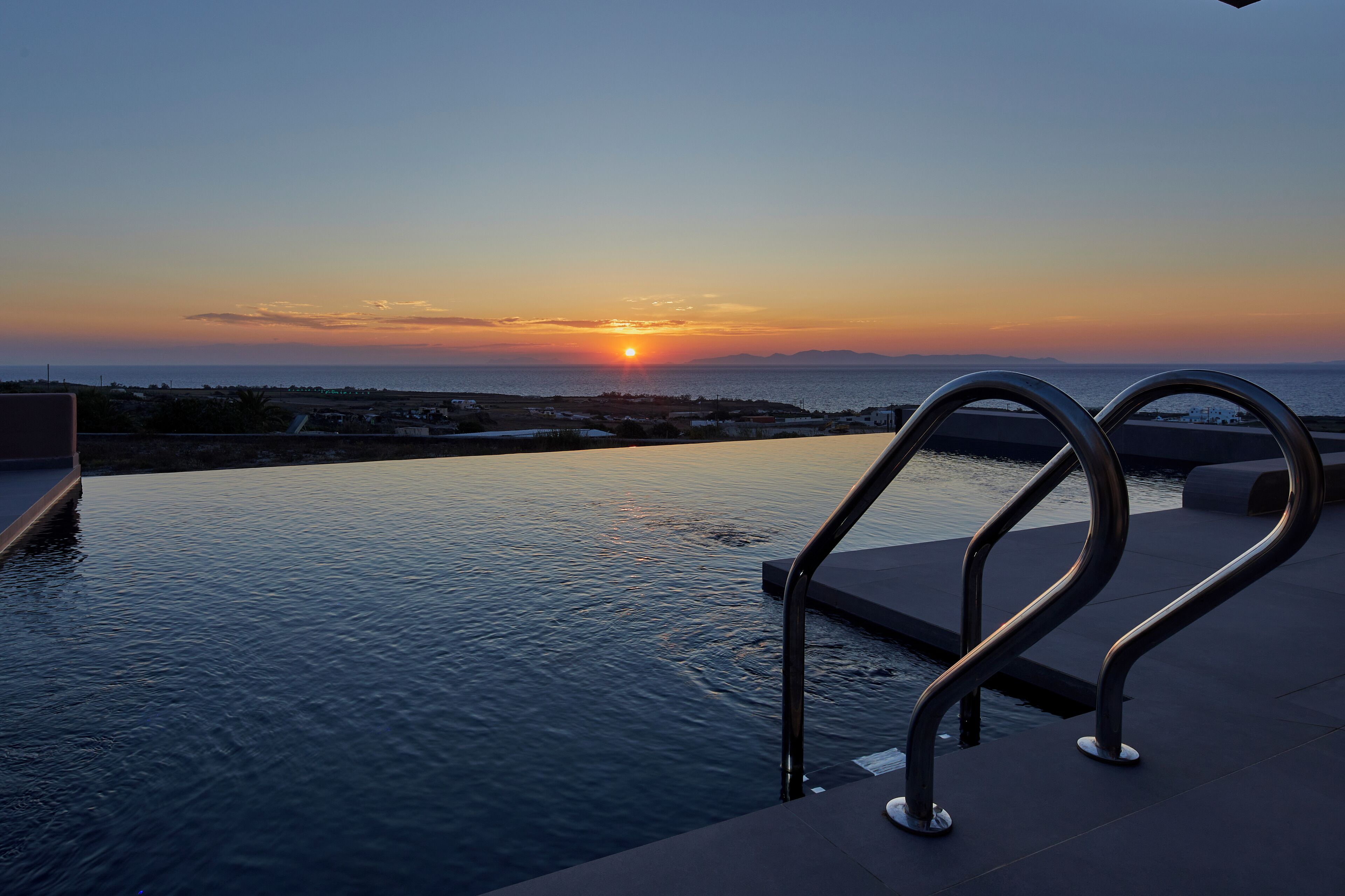 Una piscina infinita con barandillas metálicas que ofrece vistas a una puesta de sol sobre el mar.