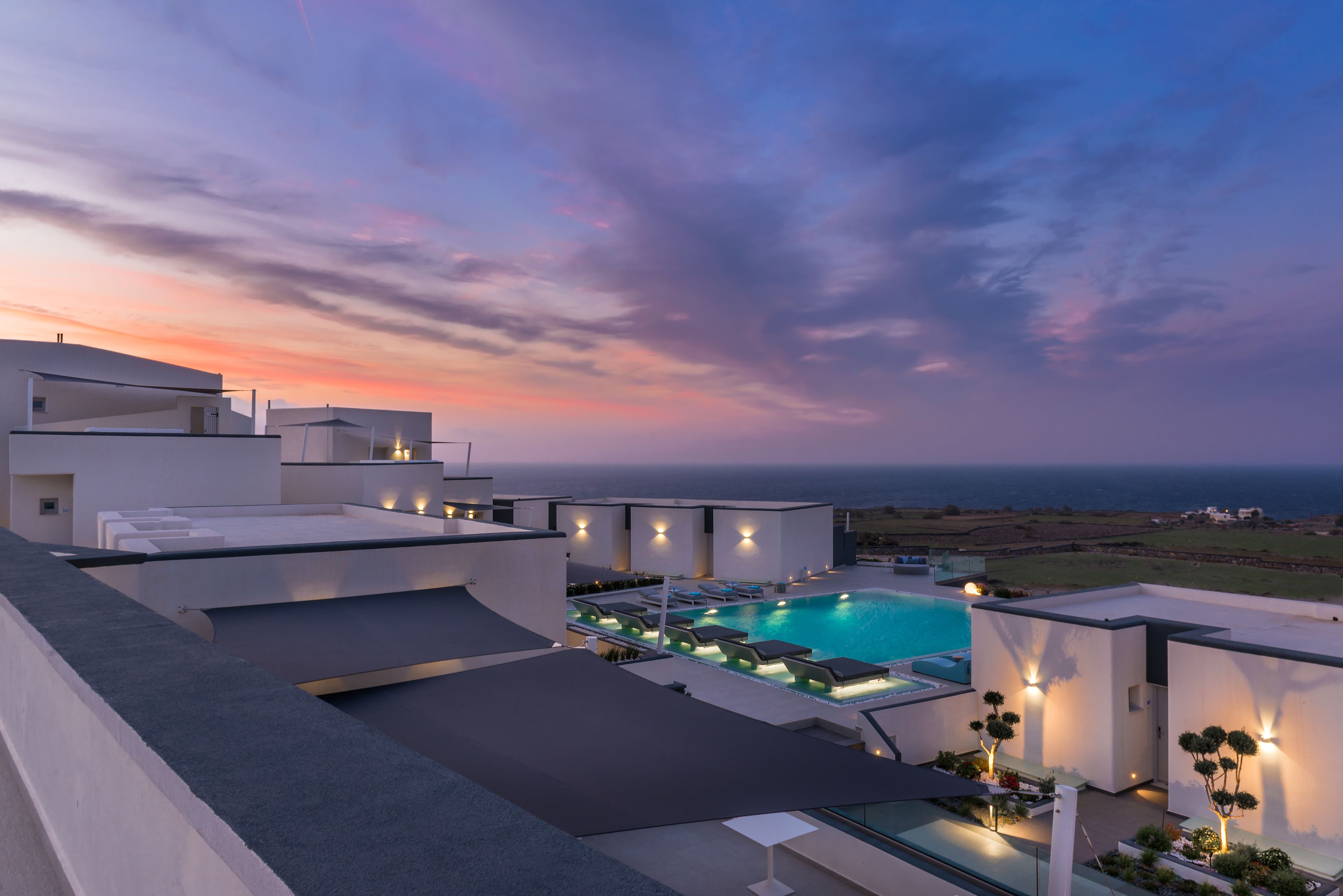 Terraço moderno de hotel com piscina, cadeiras de descanso e vista para o oceano ao pôr do sol.