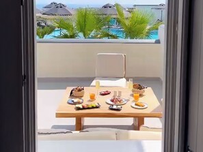 A dining table with breakfast items on a balcony overlooking a pool and palm trees. - Myst Boutique Hotel (Santorini)