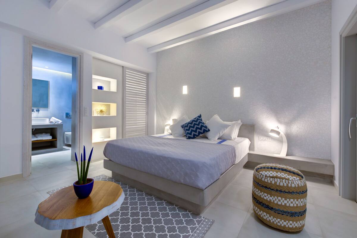 A modern bedroom with a large bed, bedside tables, a small round ottoman, and a bathroom visible through an open door.