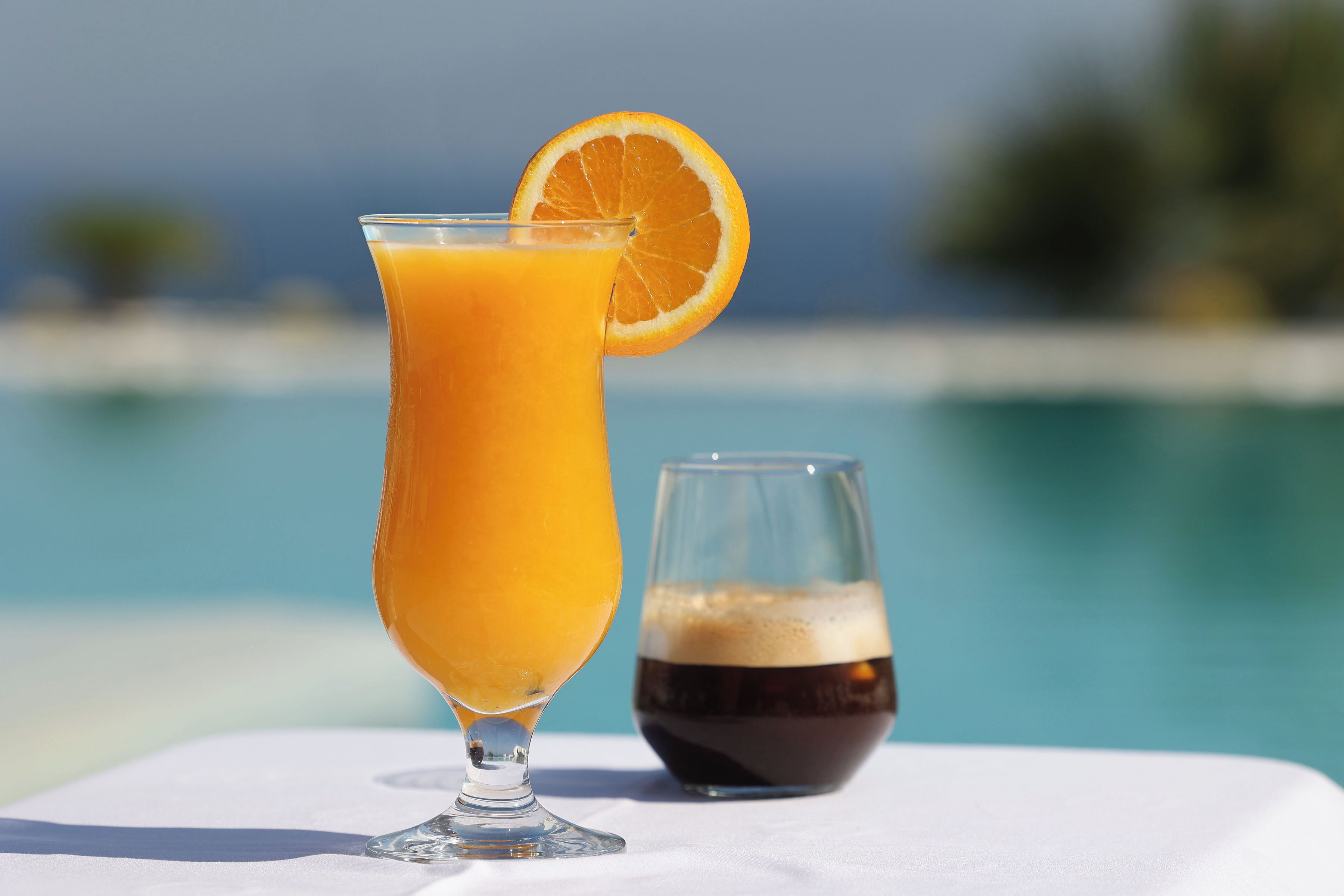 A glass of orange juice with a slice of orange on the rim, next to a glass of dark liquid, possibly coffee or tea, on a white surface by a pool.