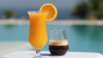A glass of orange juice with a slice of orange on the rim, next to a glass of dark liquid, possibly coffee or tea, on a white surface by a pool.