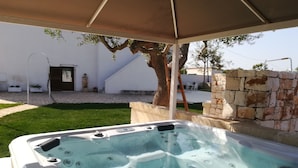 A hot tub under a canopy, surrounded by a stone wall, a tree, and a white building with a door and steps. - Masseria Poli (Conversano)