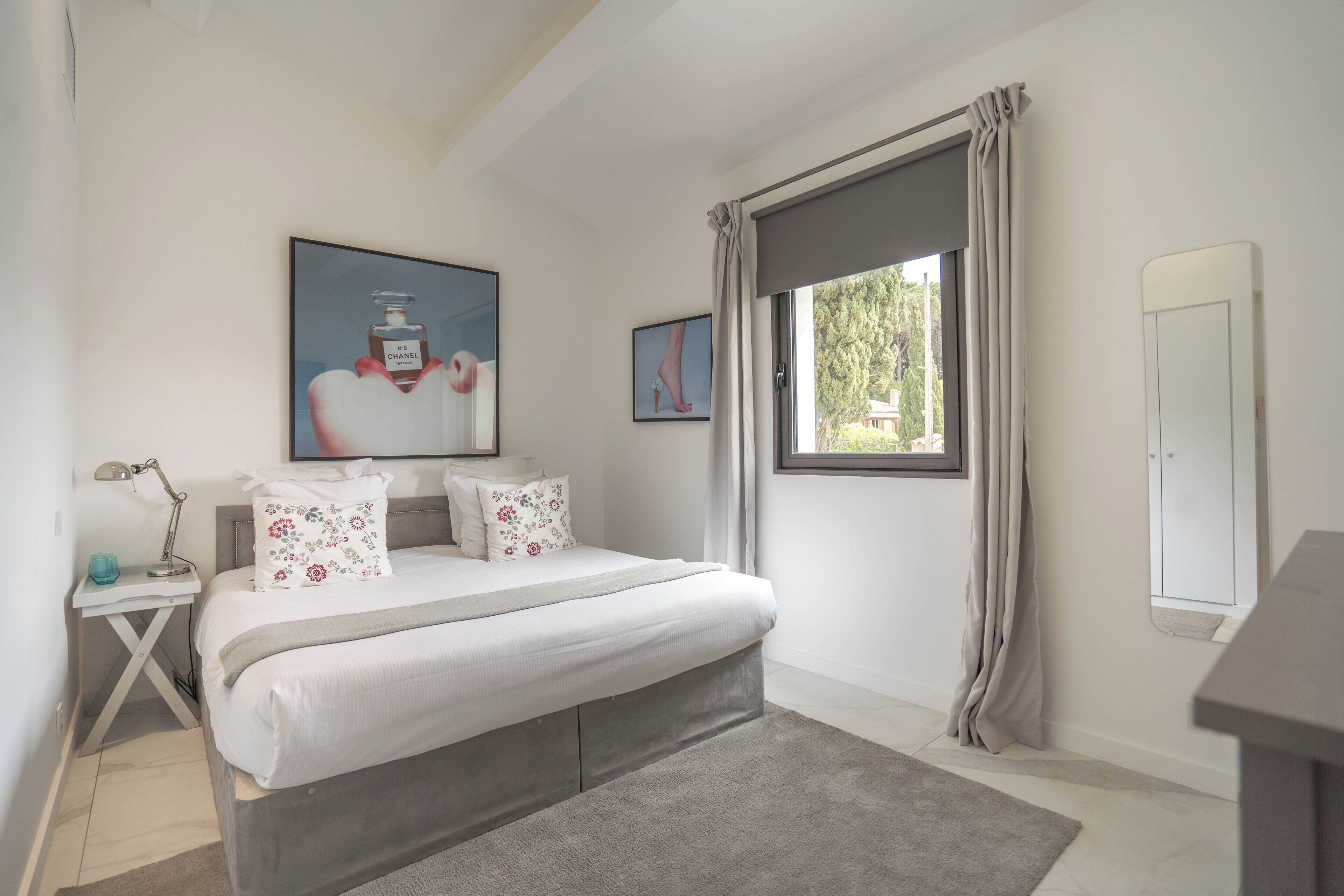 A bedroom with a bed, a nightstand, a lamp, a window with curtains, and a wall-mounted mirror.
