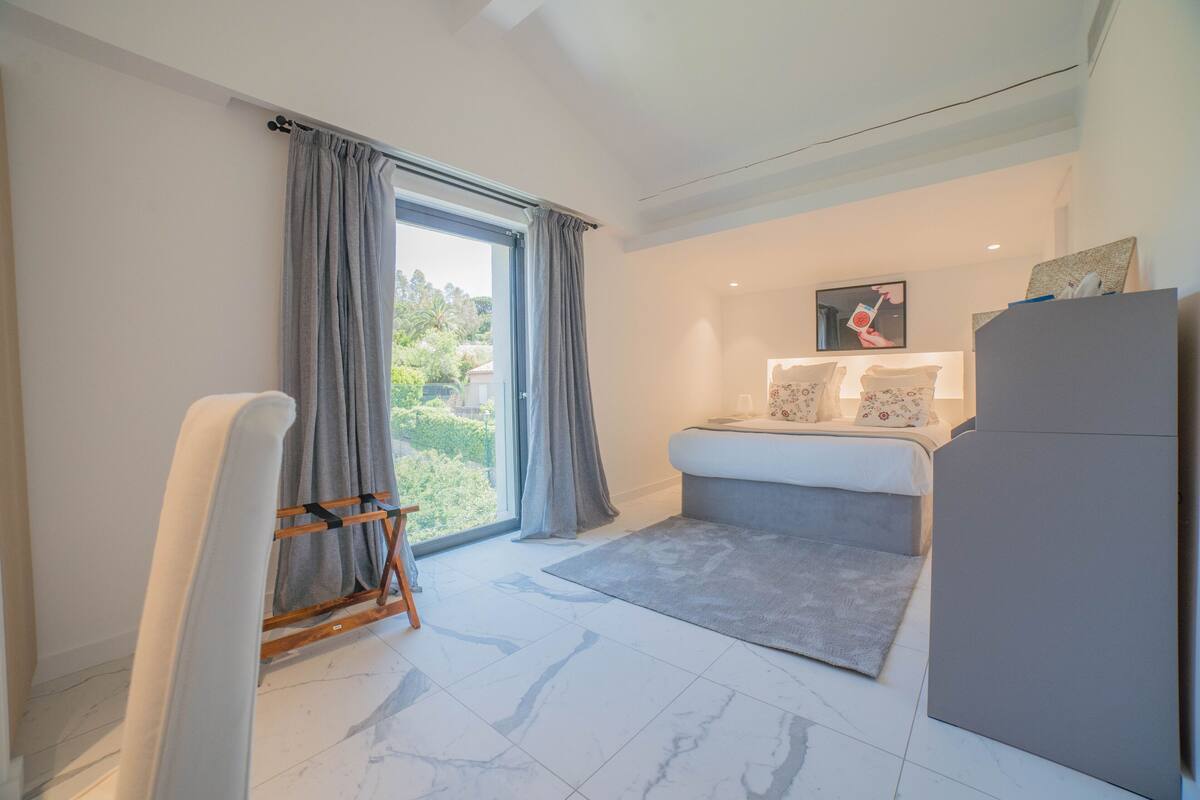 Un dormitorio moderno con un ventanal grande, una cama con cabecera gris, un sofá gris y un armario gris.