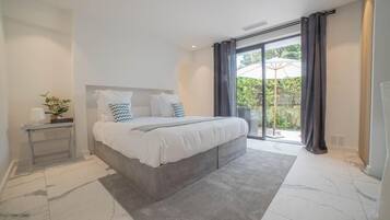A modern bedroom with a large bed, a bedside table with a plant, and a view of the outdoors through a sliding glass door.