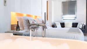 A modern hotel room with a large bed, a separate seating area, a bathtub, and a bedside table with a bottle and glasses. - Hotel Le Mandala (Saint-Tropez)