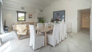 A spacious living area with a dining table, white chairs, a sofa, and a large window. - Hotel Le Mandala (Saint-Tropez)