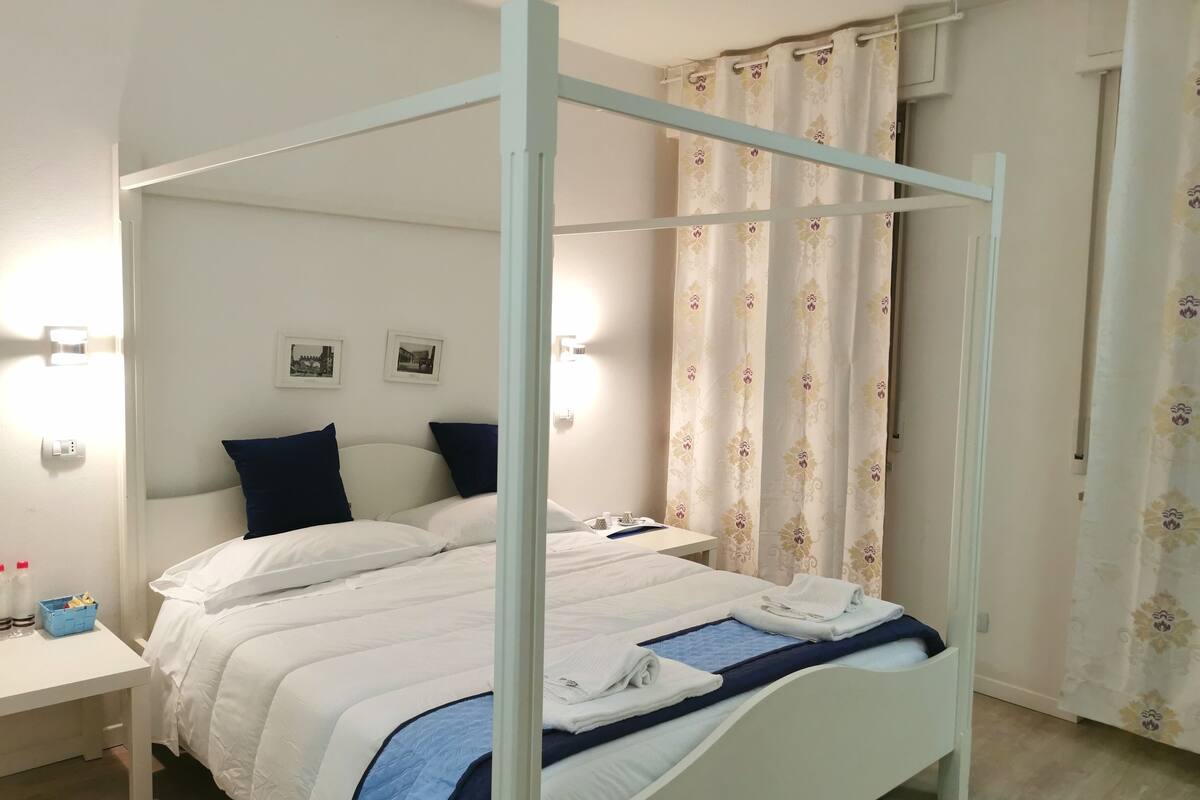 Un lit à baldaquin avec une literie blanche et des oreillers foncés, une table de chevet avec une lampe et un luminaire fixé au mur.