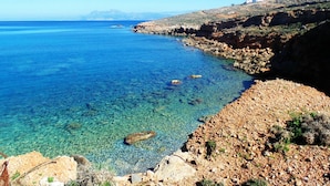 Un paysage côtier avec une mer bleue et limpide, une côte rocheuse et des montagnes à l’horizon.