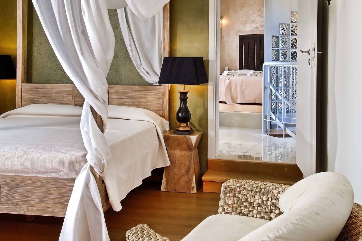 Ein Schlafzimmer mit Himmelbett, einem Korbstuhl und Blick in einen anderen Raum.