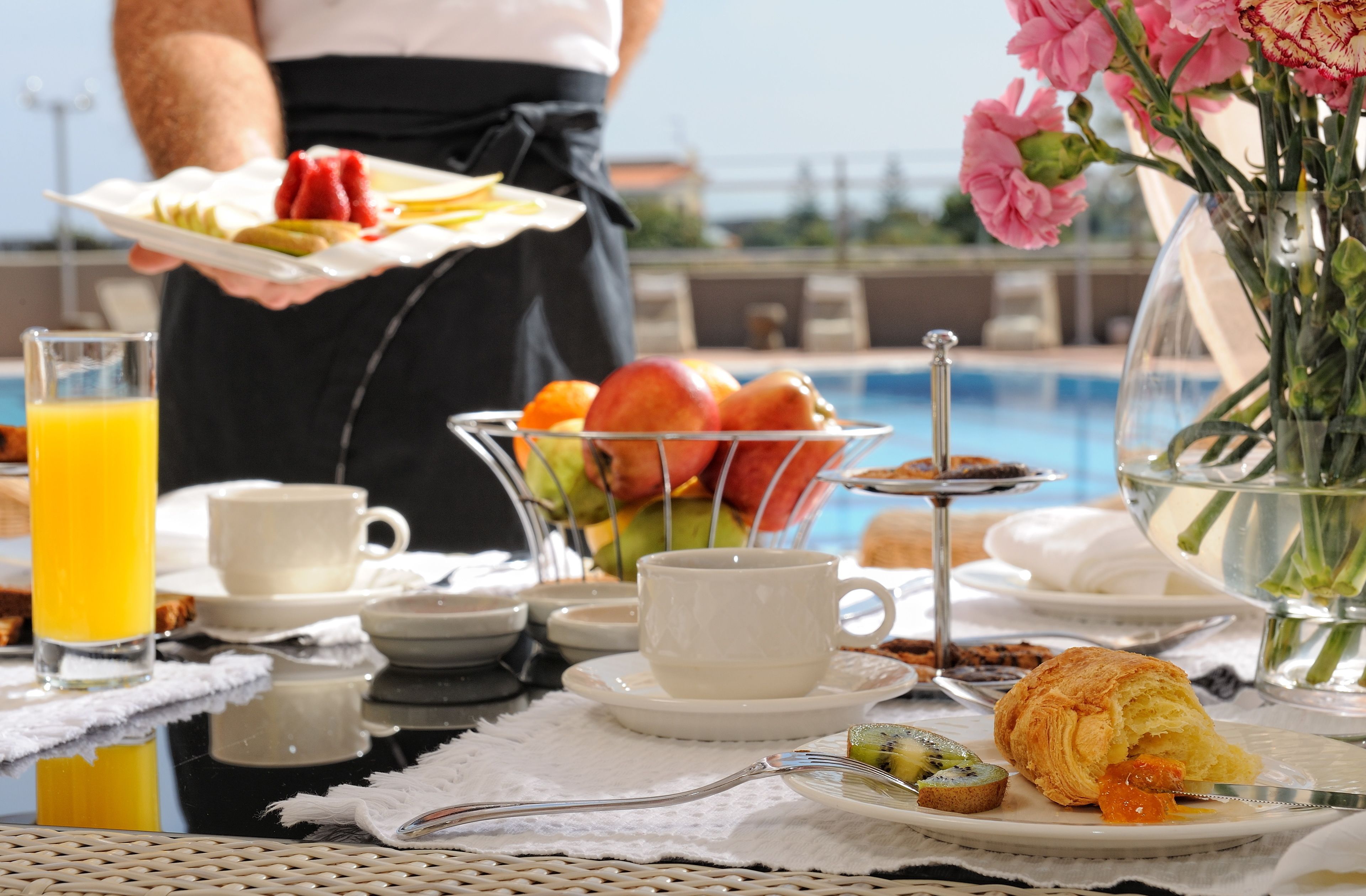 A breakfast setting with a fruit bowl, croissant, and coffee cups on a table by a pool.