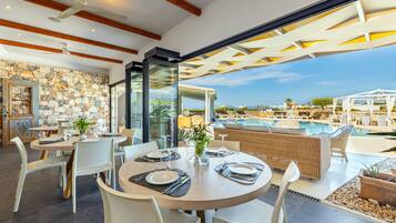 Een restaurant met ronde tafels, witte stoelen en een stenen muur. Er is uitzicht op een zwembad en terras.