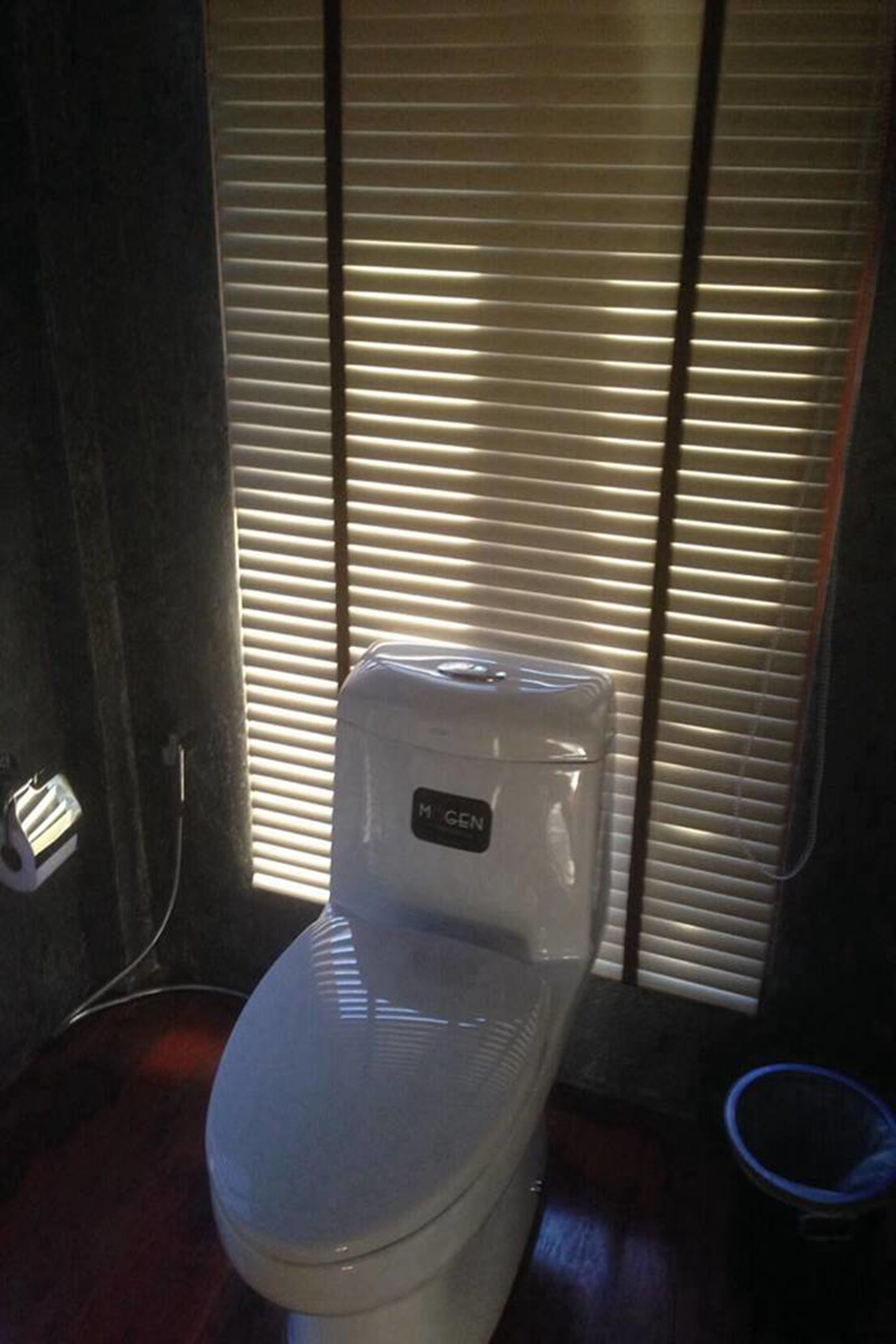A white toilet with a visible brand label in a bathroom with wooden flooring and blinds in the background.