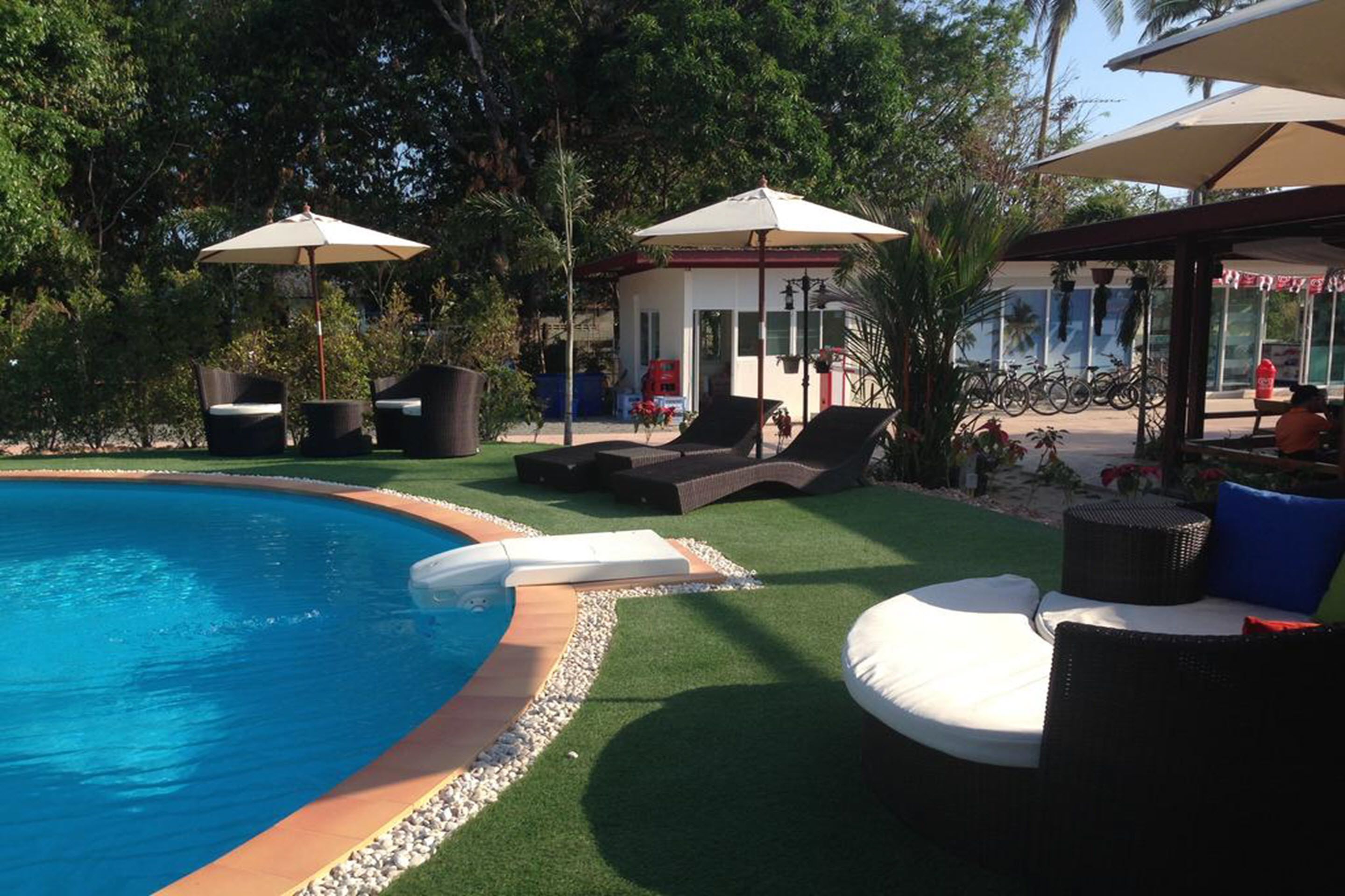 A swimming pool with lounge chairs and umbrellas, surrounded by greenery and a building with a red roof.