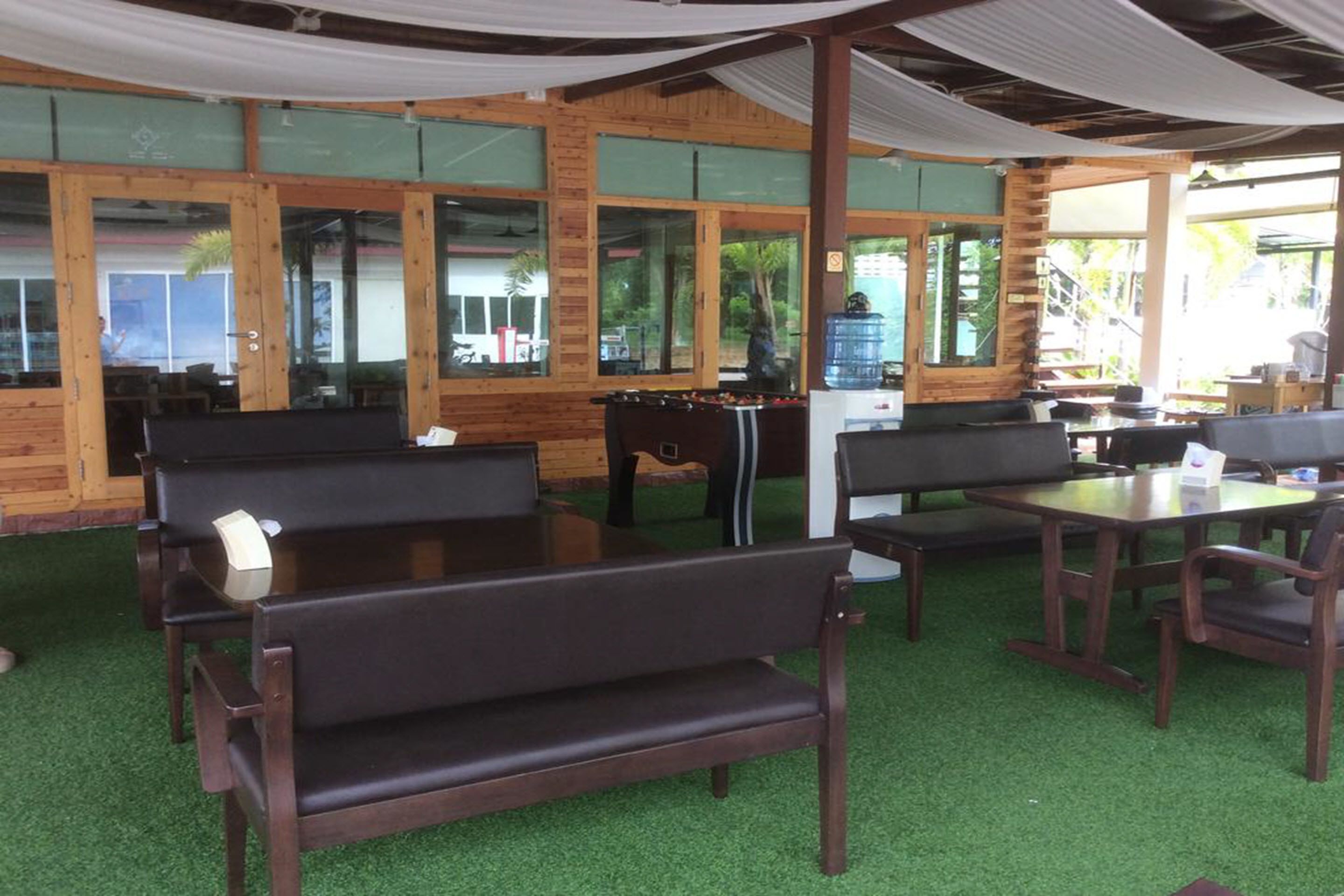 An indoor seating area with wooden tables and benches, large windows, and a green artificial turf floor.