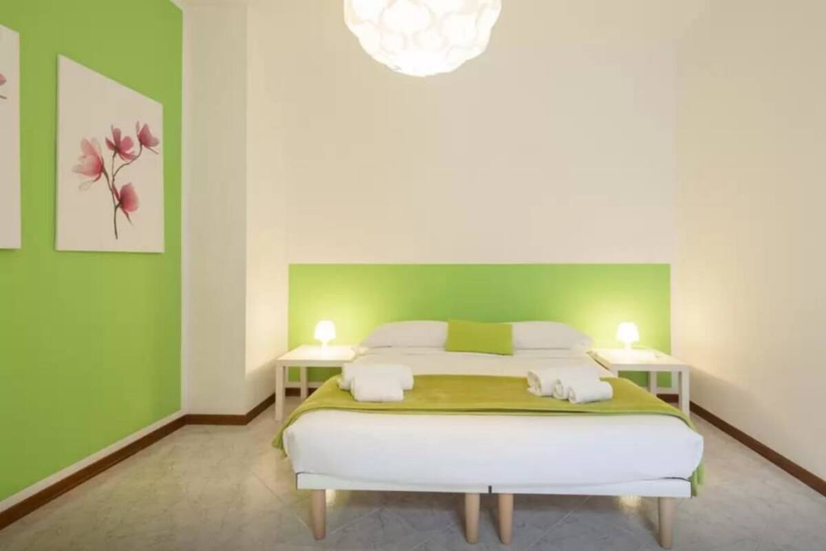 Une chambre avec un mur d’accent vert, un lit avec une literie blanche, deux tables de chevet avec des lampes et deux œuvres d’art florales encadrées accrochées au mur.