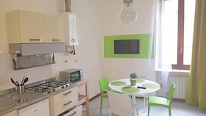Uma cozinha compacta com fogão a gás, micro-ondas e um pequeno frigorífico. Há uma mesa de jantar com cadeiras verdes e uma planta em vaso. Uma televisão de ecrã plano está fixada na parede acima da mesa.