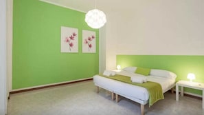 Quarto com parede verde, cama branca, um banco, duas mesas de cabeceira e um lustre.