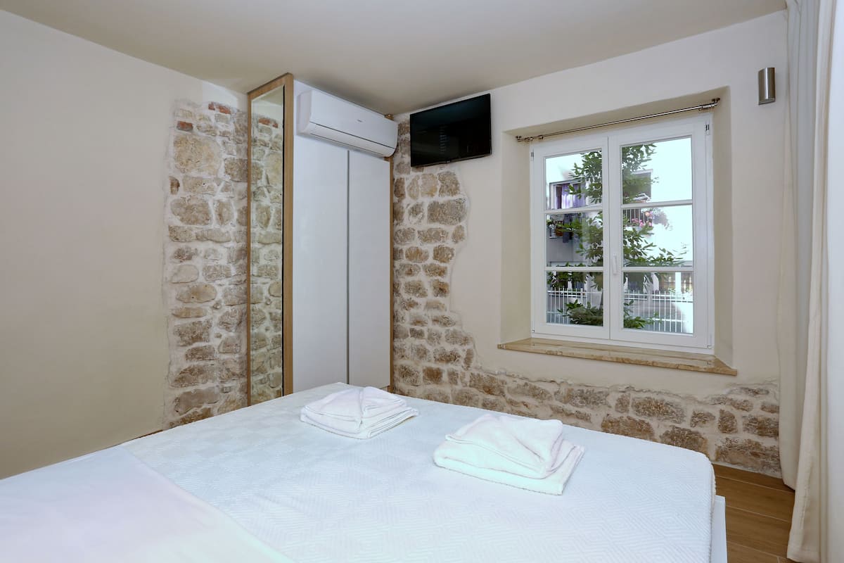 Un dormitorio con pared de piedra, una cama con sábanas blancas, un televisor montado en la pared y una ventana con vistas al follaje exterior.