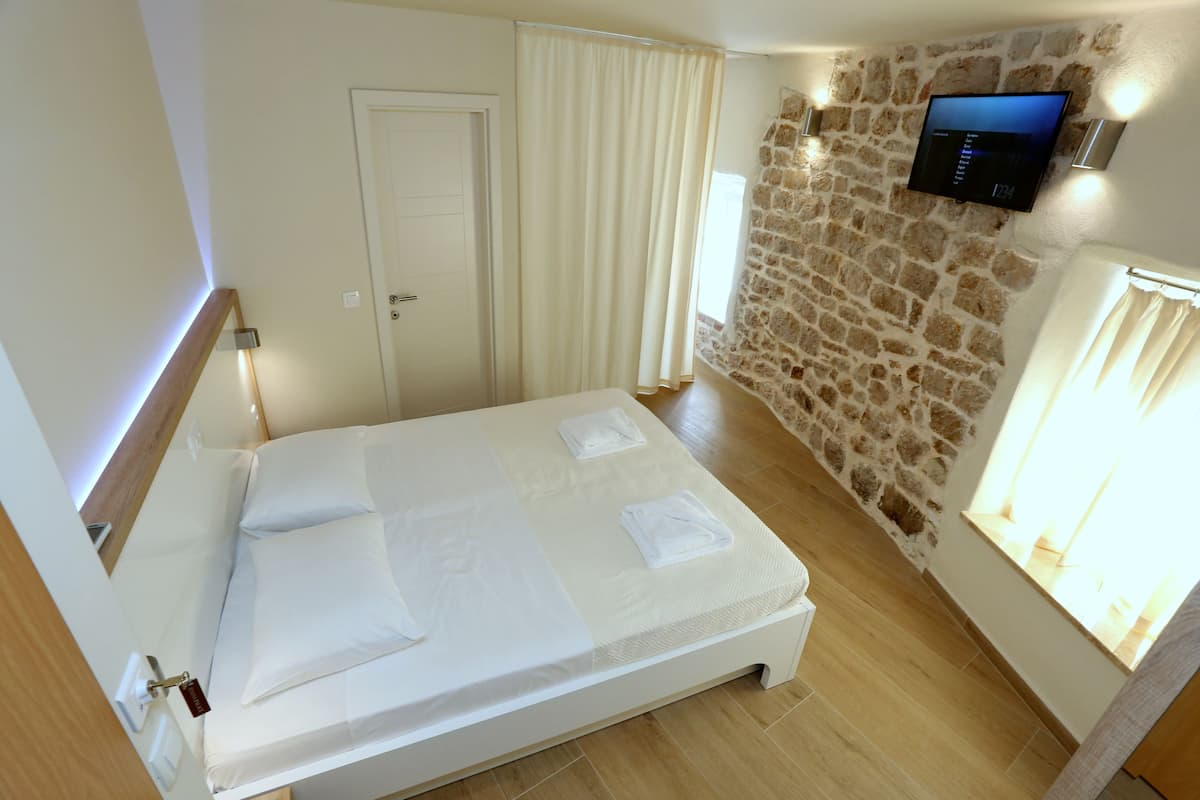 Una cama bien tendida con ropa de cama blanca, una pared de piedra, un televisor de pantalla plana montado encima y una ventana con cortinas.