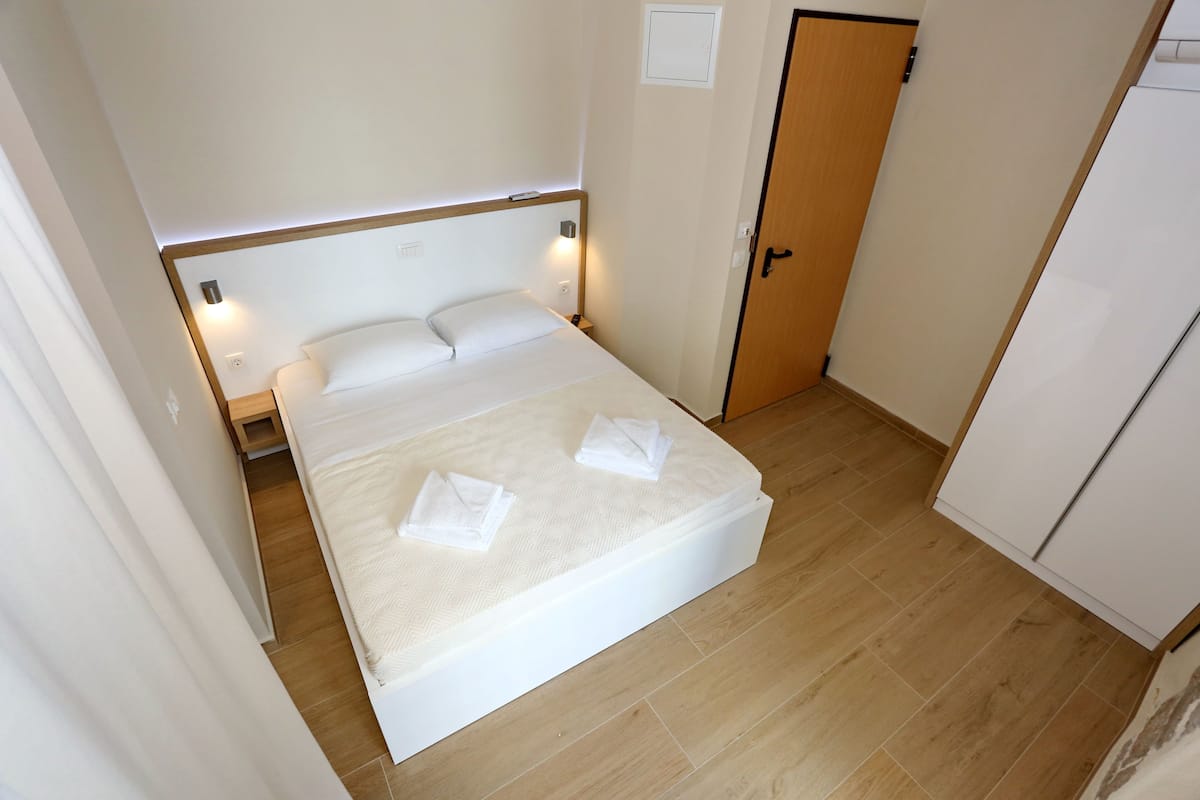 Una cama bien tendida con almohadas y toallas, una mesita de noche con lámpara, y una puerta de madera.