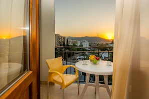 A balcony with a table and chairs, a bowl of fruit, and a cityscape view at sunset.