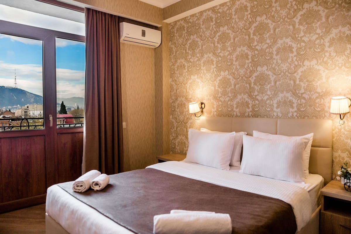 A hotel room with a large bed, two bedside lamps, a view of the city through a balcony, and an air conditioning unit.