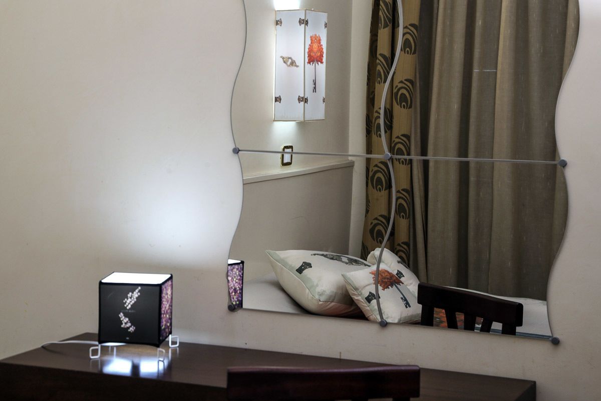 A room with a bed, a bedside table, a nightstand, a mirror, a window with curtains, and a wall-mounted shelf.