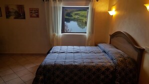Un dormitorio con cama, ventana con vista a un lago y una lámpara fijada a la pared.
