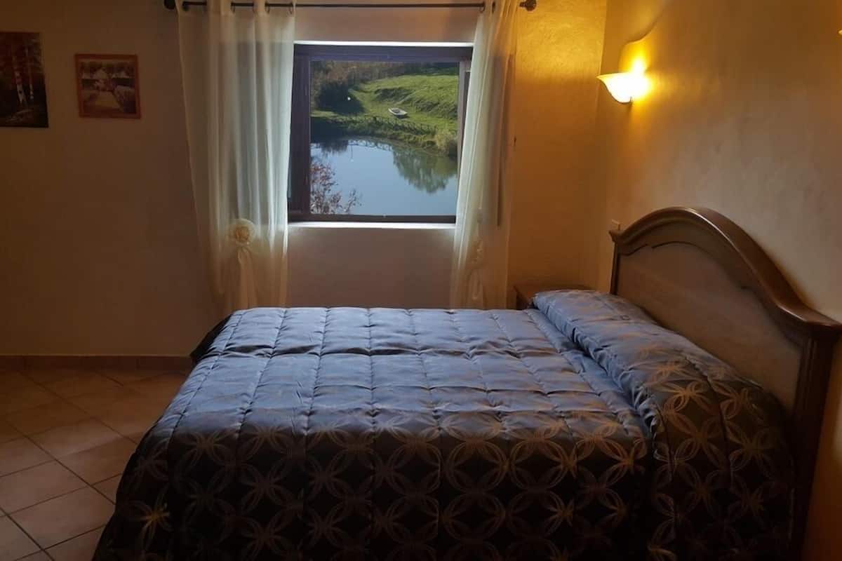 Un dormitorio con cama, ventana con vista a un lago y una lámpara fijada a la pared.