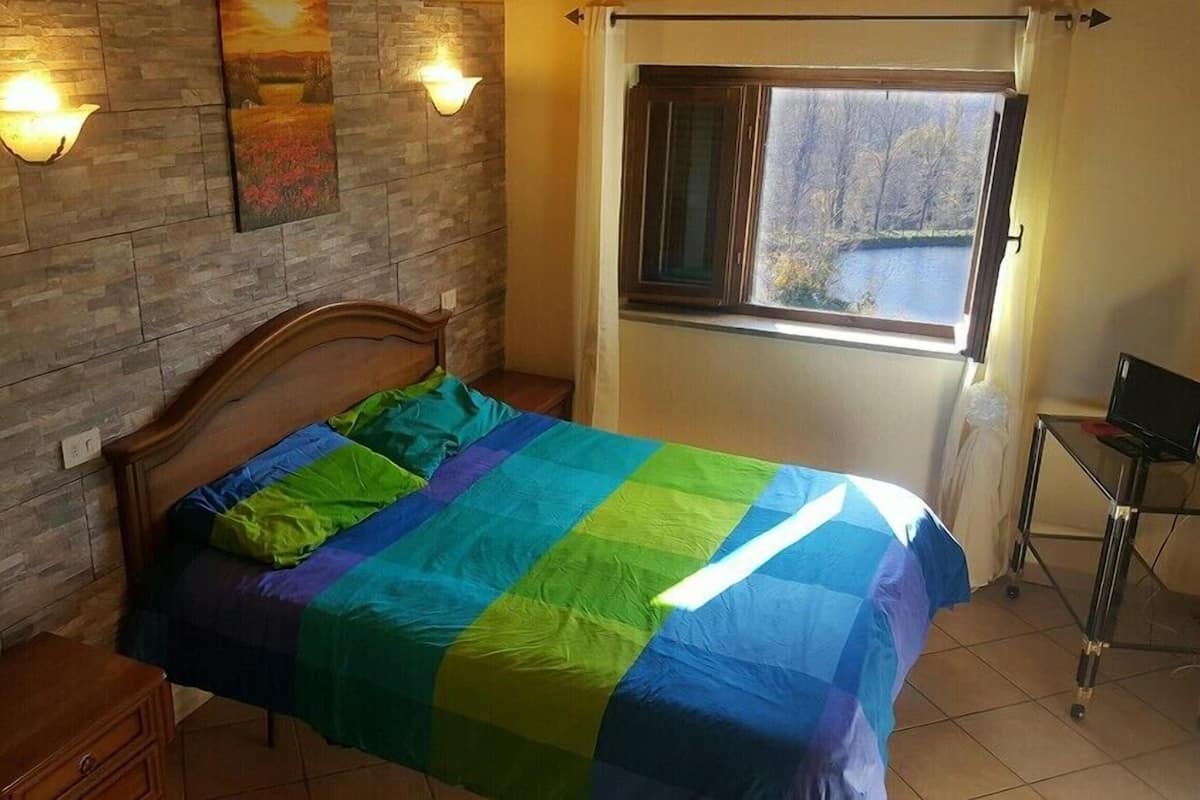 Un dormitorio con cabecera de madera, colcha colorida, mesita de noche, escritorio con computadora y ventana con vista a árboles y una masa de agua.