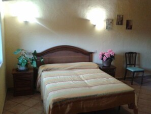 Un dormitorio con una cama de madera, una mesita de noche con una planta, una silla y un aplique de pared.