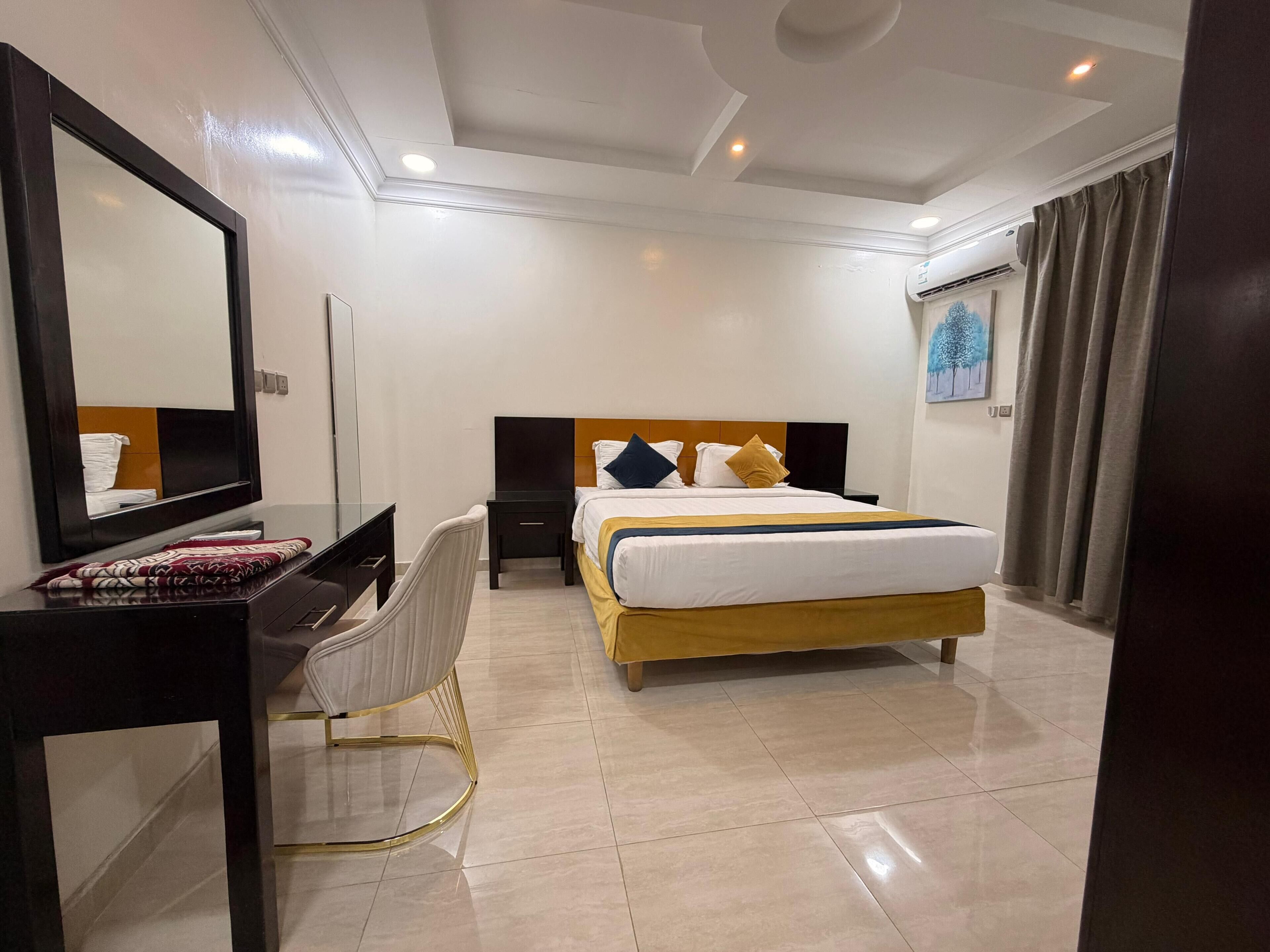 A hotel room with a bed, a desk, a chair, a mirror, and an air conditioning unit.