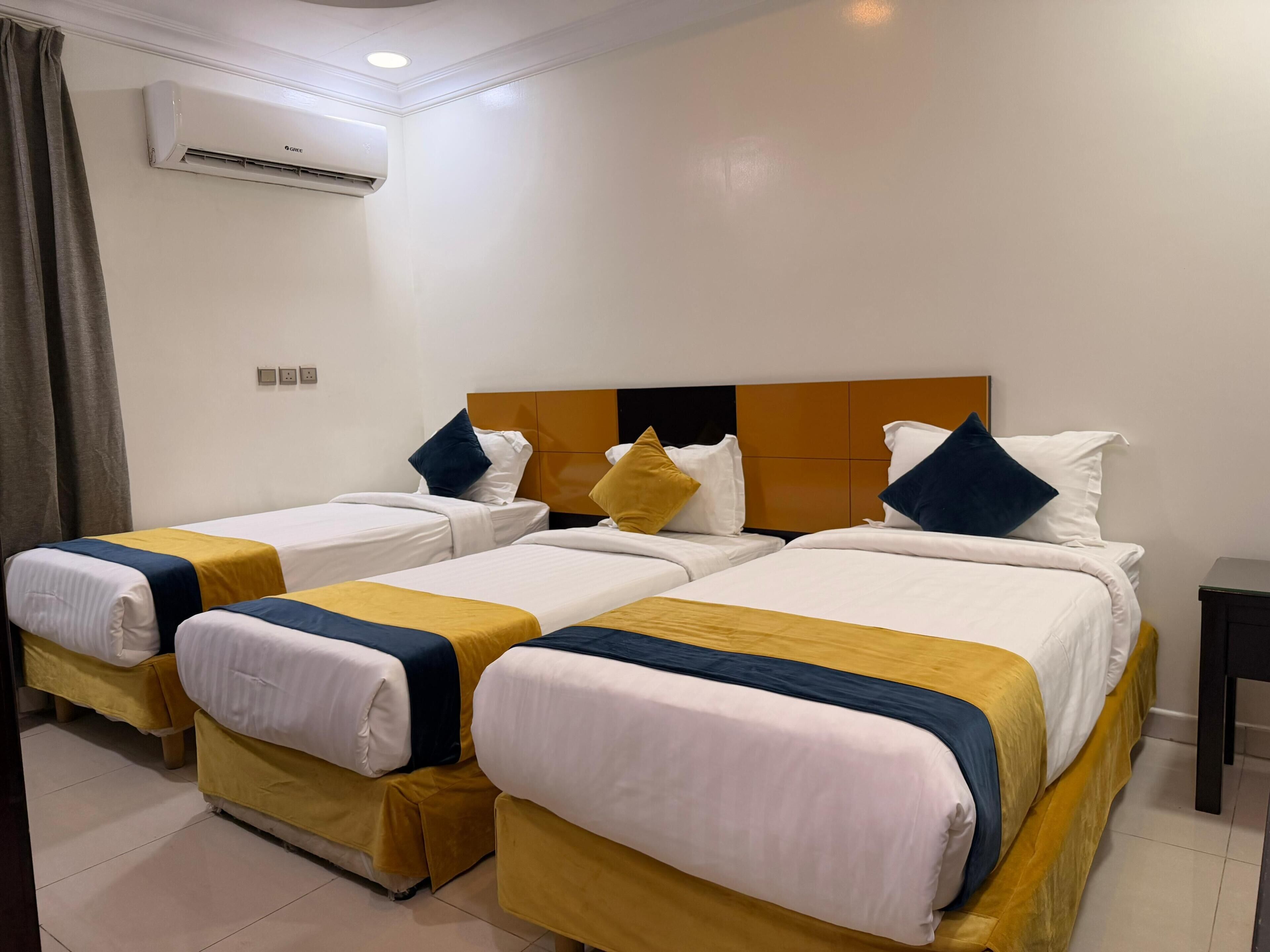 Une chambre d’hôtel avec quatre lits simples, chacun doté d’une tête de lit, de draps blancs et d’un élément décoratif jaune. Un appareil de climatisation est fixé au mur, au-dessus des lits.