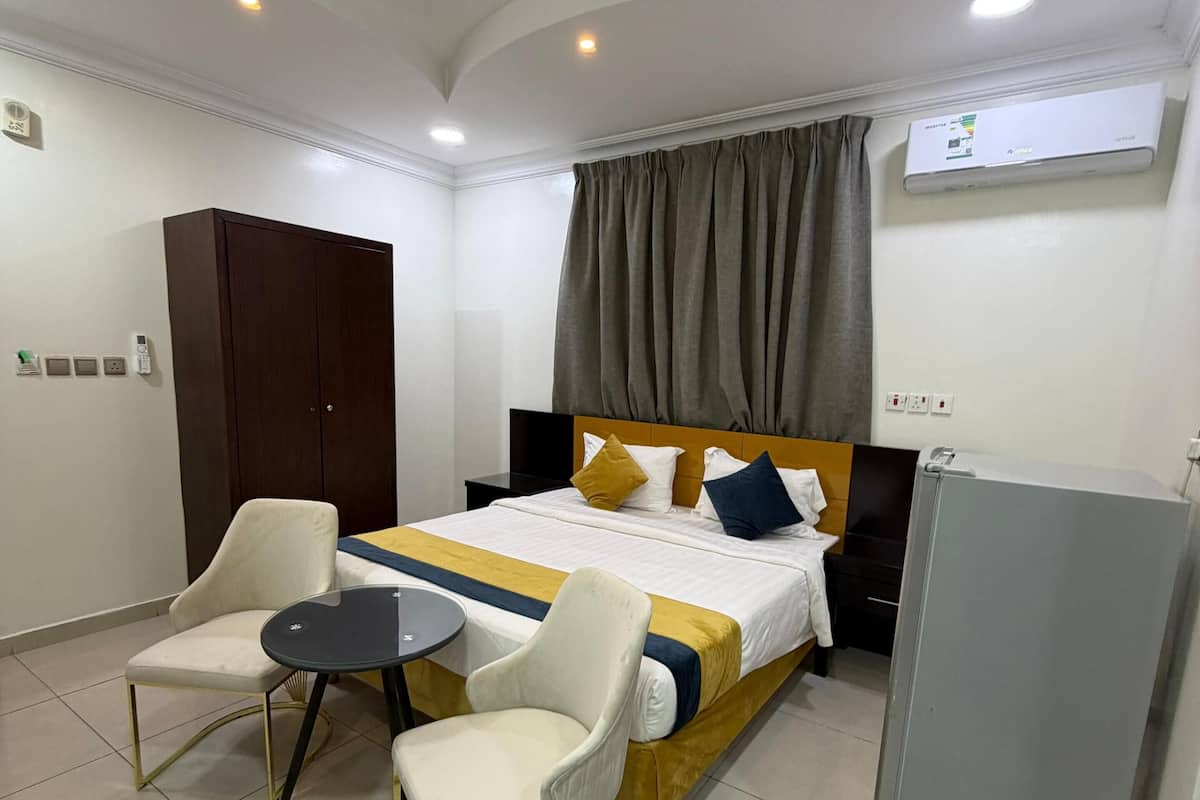 A hotel room with a bed, two chairs, a small table, a refrigerator, and an air conditioning unit.