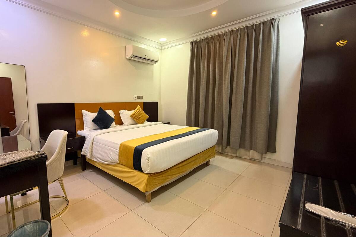 A hotel room with a bed, a desk, a chair, a mirror, an air conditioning unit, and a door.
