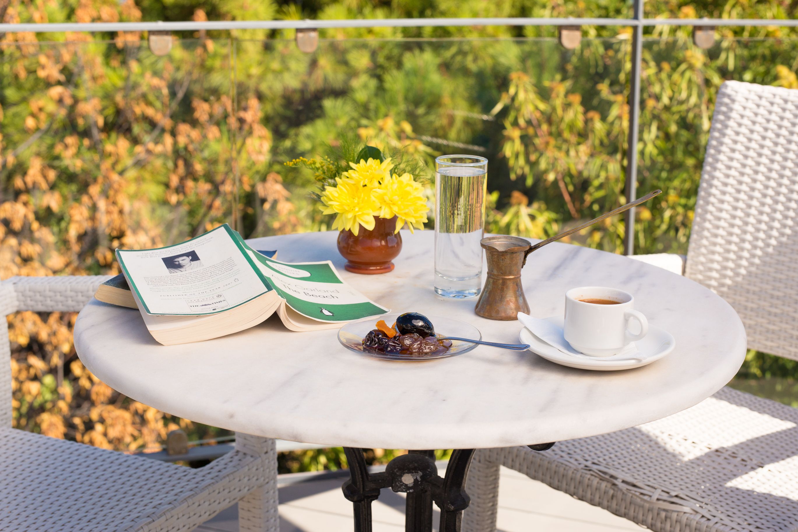 A round table with a book, a glass of water, a cup of coffee, a plate with olives, and a vase with yellow flowers.